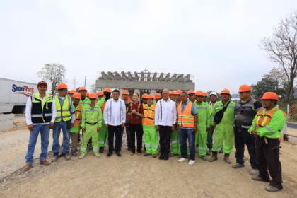 Fotografía De Plano Entero Al Aire Libre Bajo Un Cielo Nublado. En El Centro, La Presidenta Claudia Sheinbaum Pardo, Con Blusa Guinda Bordada, Y El Gobernador Javier May Rodríguez Posan Junto A Una Comitiva De Ingenieros Civiles, Mandos Y Un Numeroso Grupo De Trabajadores De La Construcción Que Visten Uniformes Fosforescentes De Color Verde Y Chalecos Reflejantes Con Cascos Naranjas. Al Fondo Se Observa La Estructura De Concreto En Construcción De Un Puente Elevado De La Carretera Macuspana–Escárcega Y Un Camión De Carga Blanco De La Empresa Supertrack.