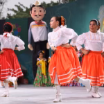 Niñas De La Compañía De Danza Folclórica De Villahermosa Bailando En El Escenario Del Teatro Al Aire Libre. Visten Blusas Blancas Con Encajes Y Faldas Naranjas Con Cintas Blancas.