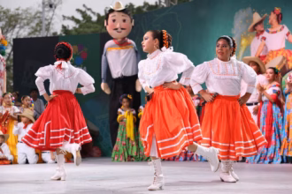Niñas De La Compañía De Danza Folclórica De Villahermosa Bailando En El Escenario Del Teatro Al Aire Libre. Visten Blusas Blancas Con Encajes Y Faldas Naranjas Con Cintas Blancas.