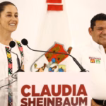 Fotografía De Plano Medio Que Muestra A La Presidenta De México, Claudia Sheinbaum Pardo, Sonriendo Detrás De Un Podio Blanco Con Su Nombre Impreso En Letras Rojas. Viste Una Blusa Blanca Con Bordados Tradicionales De Colores En Los Costados. A Su Lado Derecho Se Encuentra Javier May Rodríguez, Vistiendo Una Camisa Blanca Oficial Con Logotipos Partidistas, Ambos Listos Durante Un Acto Público.