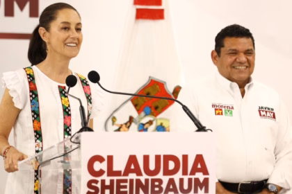 Fotografía De Plano Medio Que Muestra A La Presidenta De México, Claudia Sheinbaum Pardo, Sonriendo Detrás De Un Podio Blanco Con Su Nombre Impreso En Letras Rojas. Viste Una Blusa Blanca Con Bordados Tradicionales De Colores En Los Costados. A Su Lado Derecho Se Encuentra Javier May Rodríguez, Vistiendo Una Camisa Blanca Oficial Con Logotipos Partidistas, Ambos Listos Durante Un Acto Público.