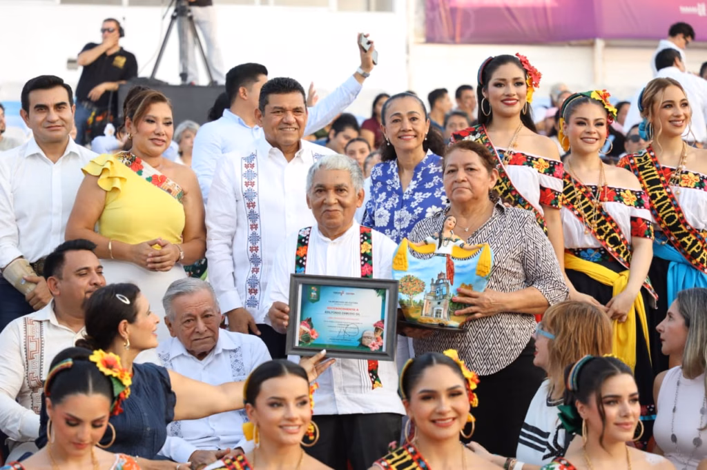Fotografía A Color Que Muestra Una Ceremonia De Premiación En Un Evento Público Al Aire Libre. En El Centro, Un Hombre De Cabello Canoso Viste Una Camisa Blanca Con Bordados Florales Y Sostiene Con Orgullo Un Reconocimiento Enmarcado.