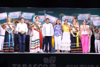 Fotografía Grupal Sobre Un Escenario Iluminado Durante La Feria Tabasco. En El Centro Destaca El Gobernador Javier May Rodríguez Con Guayabera Blanca, Acompañado Por Aurora Raleigh De La Cruz (Camisa Azul Floral) Y La Flor Tabasco 2026, María José Hernández Vera, En Traje Regional Blanco. A Los Costados Se Encuentran Las Embajadoras De Emiliano Zapata Y Cunduacán, Junto A Autoridades Municipales.