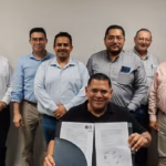 Fotografía Corporativa Horizontal En Un Espacio Cerrado De Oficinas. Al Centro, Sentado Frente A Una Mesa, El Ing. Carlos Alberto Lara Moscoso Sonríe A La Cámara Mientras Sostiene Abierto El Documento Oficial De Toma De Protesta Firmado. Detrás De Él, Una Fila De Nueve Ingenieros Y Empresarios, Miembros De La Asociación, Posan De Pie En Apoyo A La Nueva Dirigencia. A La Derecha, Se Aprecian Cuadros Con Fotografías De Expresidentes Sobre Un Muro De Madera.