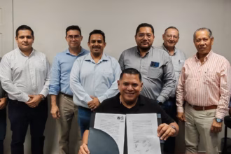 Fotografía Corporativa Horizontal En Un Espacio Cerrado De Oficinas. Al Centro, Sentado Frente A Una Mesa, El Ing. Carlos Alberto Lara Moscoso Sonríe A La Cámara Mientras Sostiene Abierto El Documento Oficial De Toma De Protesta Firmado. Detrás De Él, Una Fila De Nueve Ingenieros Y Empresarios, Miembros De La Asociación, Posan De Pie En Apoyo A La Nueva Dirigencia. A La Derecha, Se Aprecian Cuadros Con Fotografías De Expresidentes Sobre Un Muro De Madera.