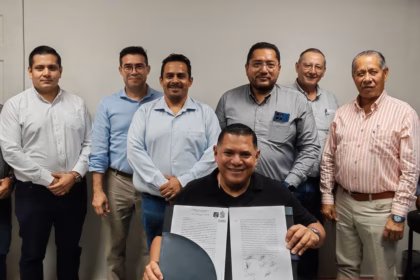 Fotografía Corporativa Horizontal En Un Espacio Cerrado De Oficinas. Al Centro, Sentado Frente A Una Mesa, El Ing. Carlos Alberto Lara Moscoso Sonríe A La Cámara Mientras Sostiene Abierto El Documento Oficial De Toma De Protesta Firmado. Detrás De Él, Una Fila De Nueve Ingenieros Y Empresarios, Miembros De La Asociación, Posan De Pie En Apoyo A La Nueva Dirigencia. A La Derecha, Se Aprecian Cuadros Con Fotografías De Expresidentes Sobre Un Muro De Madera.