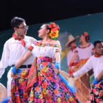 Una Pareja De Bailarines De Folclor Tabasqueño Posa Sonriente Durante Una Presentación En La Feria Tabasco 2026. La Mujer Viste Una Falda Floreada De Colores Vibrantes Y Flores En El Cabello, Mientras El Hombre Usa Una Camisa Blanca, Paliacate Rojo Y Sostiene Un Sombrero.