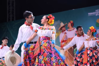 Una Pareja De Bailarines De Folclor Tabasqueño Posa Sonriente Durante Una Presentación En La Feria Tabasco 2026. La Mujer Viste Una Falda Floreada De Colores Vibrantes Y Flores En El Cabello, Mientras El Hombre Usa Una Camisa Blanca, Paliacate Rojo Y Sostiene Un Sombrero.