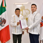 Fotografía Institucional En Interiores Con Iluminación Clara Y Plano Medio. Dos Hombres Se Encuentran De Pie, Posicionados De Frente Hacia La Cámara Mientras Sostienen Conjuntamente Un Documento Oficial Con Carpeta De Bordes Guinda. El Hombre De La Izquierda, El Gobernador Javier May Rodríguez, Viste Una Guayabera Blanca Formal De Manga Larga Con Detalles Bordados Verticales En Tonos Claros Y Pantalones Oscuros. El Hombre De La Derecha, El Comisario Alejandro Leal López, Viste Una Guayabera Blanca Similar Con Bordados Geométricos Coloridos En Los Hombros Y El Pecho, Combinada Con Pantalones Oscuros. Flanqueando A Ambos Personajes, Se Ubica La Bandera De México En El Extremo Izquierdo Y La Bandera Del Estado De Tabasco En El Extremo Derecho. Al Fondo, Sobre La Pared Blanca, Resalta Un Escudo De Armas Dorado Montado En Una Placa De Vidrio Transparente.