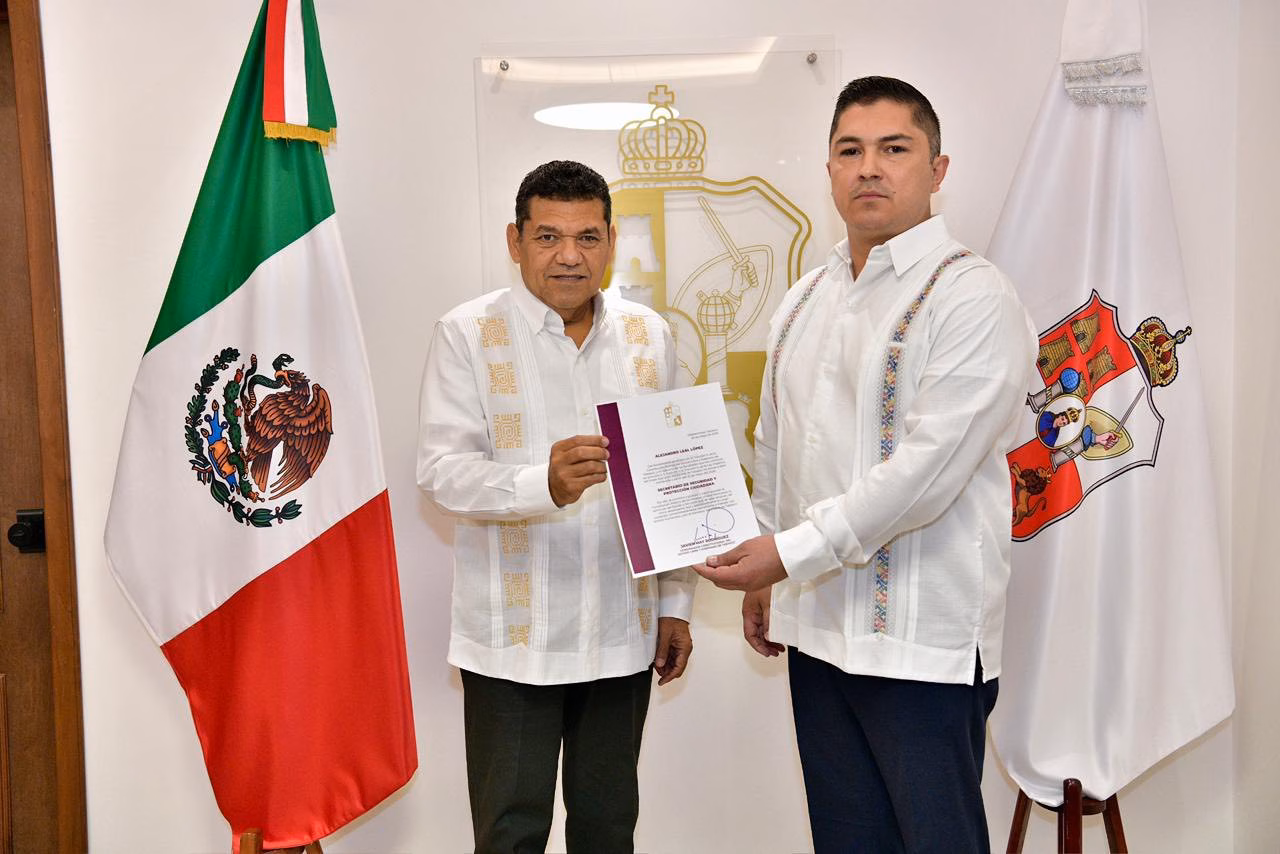 Gobernador Javier May Designa Al Comisario Federal Alejandro Leal López Como Nuevo Titular De La Sspc En Tabasco 70 Fotografía Institucional En Interiores Con Iluminación Clara Y Plano Medio. Dos Hombres Se Encuentran De Pie, Posicionados De Frente Hacia La Cámara Mientras Sostienen Conjuntamente Un Documento Oficial Con Carpeta De Bordes Guinda. El Hombre De La Izquierda, El Gobernador Javier May Rodríguez, Viste Una Guayabera Blanca Formal De Manga Larga Con Detalles Bordados Verticales En Tonos Claros Y Pantalones Oscuros. El Hombre De La Derecha, El Comisario Alejandro Leal López, Viste Una Guayabera Blanca Similar Con Bordados Geométricos Coloridos En Los Hombros Y El Pecho, Combinada Con Pantalones Oscuros. Flanqueando A Ambos Personajes, Se Ubica La Bandera De México En El Extremo Izquierdo Y La Bandera Del Estado De Tabasco En El Extremo Derecho. Al Fondo, Sobre La Pared Blanca, Resalta Un Escudo De Armas Dorado Montado En Una Placa De Vidrio Transparente.