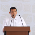 Primer Plano Del Gobernador Javier May Rodríguez Durante Una Rueda De Prensa. Se Encuentra Hablando Frente A Un Micrófono En Un Podio De Madera Que Muestra El Escudo Oficial De Tabasco.
