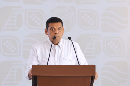 Primer Plano Del Gobernador Javier May Rodríguez Durante Una Rueda De Prensa. Se Encuentra Hablando Frente A Un Micrófono En Un Podio De Madera Que Muestra El Escudo Oficial De Tabasco.