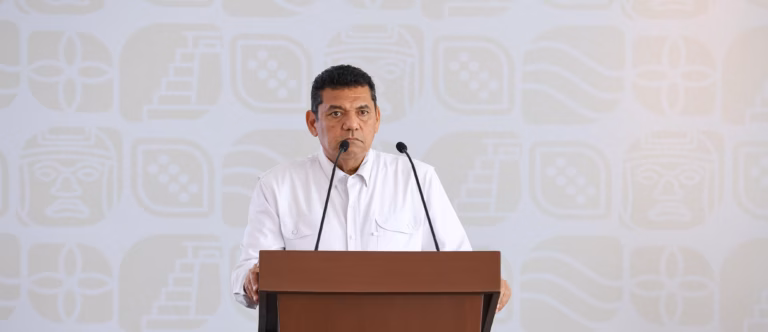 Primer Plano Del Gobernador Javier May Rodríguez Durante Una Rueda De Prensa. Se Encuentra Hablando Frente A Un Micrófono En Un Podio De Madera Que Muestra El Escudo Oficial De Tabasco.