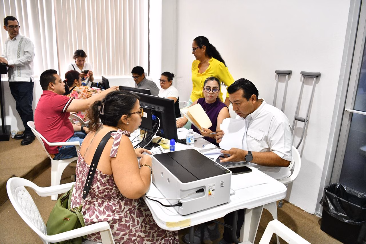 Fotografía En Plano Medio De Una Mesa De Atención Administrativa Donde Personal Del Gobierno Del Estado Revisa Documentos De Ciudadanos. En Primer Plano, Un Hombre Con Camisa Blanca De Manga Corta Y Una Mujer Con Blusa Morada Analizan Expedientes En Papel Y Consultan Una Computadora De Escritorio.