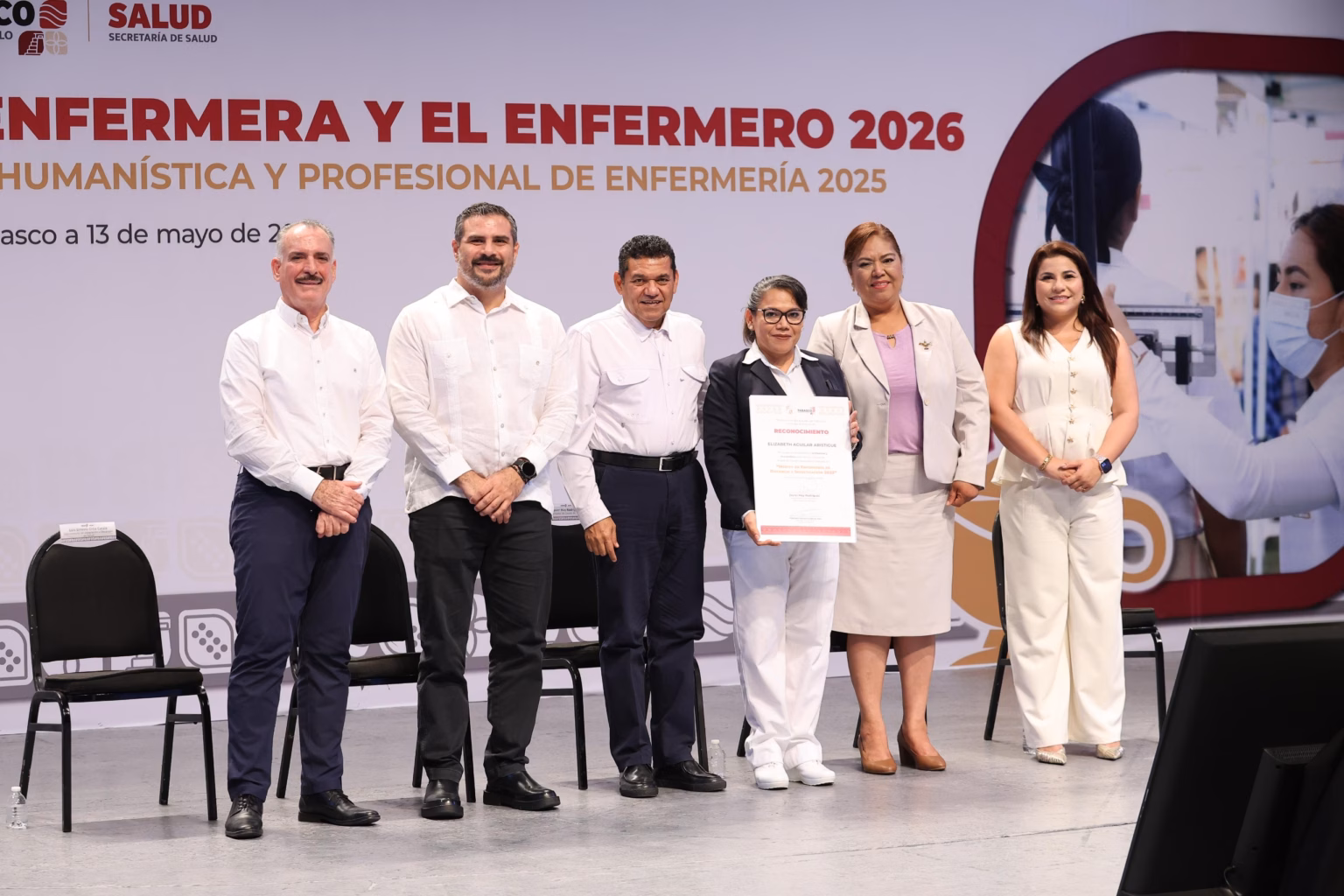 Javier May Reconoce Labor De Enfermeras Y Enfermeros En Tabasco 69 El Gobernador De Tabasco, Javier May Rodríguez, Entrega Un Reconocimiento Impreso A Una Profesional De La Enfermería En El Escenario Del Teatro Esperanza Iris. El Mandatario Viste Una Guayabera Blanca Tradicional Y Sonríe Mientras Sostiene El Galardón Junto A La Enfermera. Al Fondo Se Observa El Logotipo Oficial Del Gobierno De Tabasco Y Un Arreglo Floral.