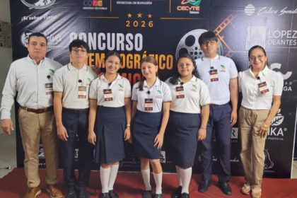 Otografía Horizontal En Plano General Que Captura La Ceremonia De Inauguración Del Concurso Cinematográfico Académico En Un Auditorio. En La Parte Izquierda, Un Hombre Con Camisa Blanca De Manga Larga Y Pantalón Caqui Permanece De Pie Junto A Un Grupo De Cinco Estudiantes (Tres Mujeres Y Dos Hombres) Alineados Al Centro; Todos Los Alumnos Visten El Uniforme Institucional Conformado Por Camisas Polo Blancas Con Cuellos Oscuros, Pantalones O Faldas Oscuras Y Zapatos Negros.