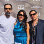 Plano Medio De Tres Personas Posando Sonrientes De Pie Frente A Un Fondo Gris Texturizado Con Los Logotipos Repetidos De 'Kurdistan Film Commission' Y 'Marché Du Film'. A La Izquierda, Un Hombre Con Barba Encanecida Y Gafas De Sol Viste Una Playera Gris Claro De Manga Corta. Al Centro, La Actriz Mexicana Alejandra De La O Sonríe Usando Gafas De Sol Oscuras, Aretes Largos Y Un Elegante Top Satinado De Cuello Halter En Color Azul Turquesa Con Un Lazo En La Cintura.