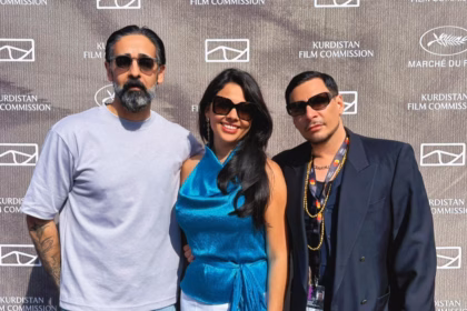 Plano Medio De Tres Personas Posando Sonrientes De Pie Frente A Un Fondo Gris Texturizado Con Los Logotipos Repetidos De 'Kurdistan Film Commission' Y 'Marché Du Film'. A La Izquierda, Un Hombre Con Barba Encanecida Y Gafas De Sol Viste Una Playera Gris Claro De Manga Corta. Al Centro, La Actriz Mexicana Alejandra De La O Sonríe Usando Gafas De Sol Oscuras, Aretes Largos Y Un Elegante Top Satinado De Cuello Halter En Color Azul Turquesa Con Un Lazo En La Cintura.
