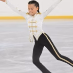 Fotografía De La Niña Patinadora Mariana Rovirosa Priego En Plena Competencia Sobre Una Pista De Hielo. Se Le Observa Deslizándose Con Los Brazos Extendidos Y El Cuerpo Inclinado Hacia Adelante En Una Postura Técnica De Patinaje Artístico. Viste Un Traje De Gala Inspirado En Michael Jackson, Compuesto Por Una Chaqueta Blanca De Cuello Alto Con Detalles Y Bordados Dorados, Y Un Pantalón Negro Con Una Franja Dorada Brillante En Los Costados. Su Cabello Está Peinado Con Trenzas Pegadas Y Al Fondo Se Aprecia El Muro Perimetral De La Pista.