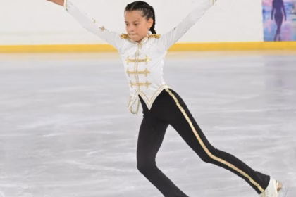 Fotografía De La Niña Patinadora Mariana Rovirosa Priego En Plena Competencia Sobre Una Pista De Hielo. Se Le Observa Deslizándose Con Los Brazos Extendidos Y El Cuerpo Inclinado Hacia Adelante En Una Postura Técnica De Patinaje Artístico. Viste Un Traje De Gala Inspirado En Michael Jackson, Compuesto Por Una Chaqueta Blanca De Cuello Alto Con Detalles Y Bordados Dorados, Y Un Pantalón Negro Con Una Franja Dorada Brillante En Los Costados. Su Cabello Está Peinado Con Trenzas Pegadas Y Al Fondo Se Aprecia El Muro Perimetral De La Pista.