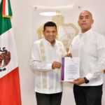 Fotografía Institucional En Interiores Con Iluminación Clara Y Plano Medio. Dos Hombres Se Encuentran De Pie Posando De Frente Hacia La Cámara Mientras Sostienen Juntos Una Carpeta Oficial Blanca Con Bordes Guinda Que Contiene Un Documento Firmado. A La Izquierda, El Gobernador Javier May Rodríguez Viste Una Guayabera Blanca Formal De Manga Larga Con Bordados Geométricos Verticales En Tonos Claros Y Pantalones Oscuros. A La Derecha, El Nuevo Comisionado De La Policía Estatal, Omar Castañeda Ríos.