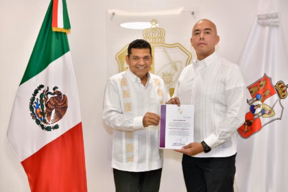 Fotografía Institucional En Interiores Con Iluminación Clara Y Plano Medio. Dos Hombres Se Encuentran De Pie Posando De Frente Hacia La Cámara Mientras Sostienen Juntos Una Carpeta Oficial Blanca Con Bordes Guinda Que Contiene Un Documento Firmado. A La Izquierda, El Gobernador Javier May Rodríguez Viste Una Guayabera Blanca Formal De Manga Larga Con Bordados Geométricos Verticales En Tonos Claros Y Pantalones Oscuros. A La Derecha, El Nuevo Comisionado De La Policía Estatal, Omar Castañeda Ríos.