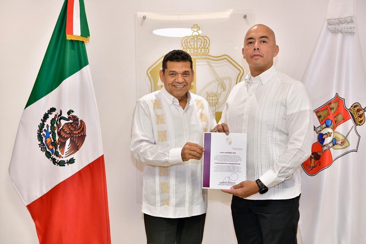 ¿Quién Es Omar Castañeda Ríos? Conoce El Perfil Del Nuevo Comisionado De La Policía Estatal De Tabasco 70 Fotografía Institucional En Interiores Con Iluminación Clara Y Plano Medio. Dos Hombres Se Encuentran De Pie Posando De Frente Hacia La Cámara Mientras Sostienen Juntos Una Carpeta Oficial Blanca Con Bordes Guinda Que Contiene Un Documento Firmado. A La Izquierda, El Gobernador Javier May Rodríguez Viste Una Guayabera Blanca Formal De Manga Larga Con Bordados Geométricos Verticales En Tonos Claros Y Pantalones Oscuros. A La Derecha, El Nuevo Comisionado De La Policía Estatal, Omar Castañeda Ríos.