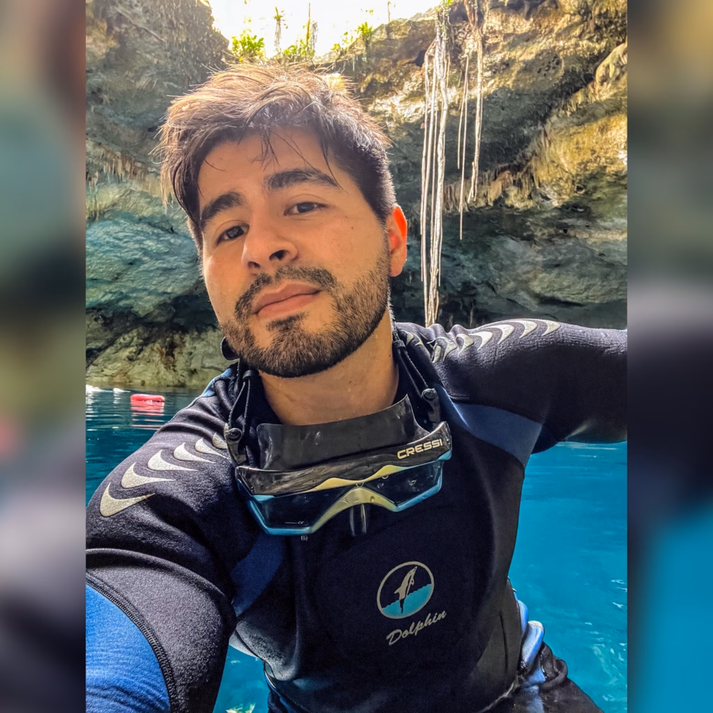 &Quot;Salvajes Sin Límites&Quot;: Lanzan Serie Documental Sobre La Biodiversidad De Los 17 Municipios De Tabasco 3 Un Hombre Joven Con Barba Corta Se Toma Una Selfi Vistiendo Un Traje De Neopreno Negro Y Azul Con Una Máscara De Buceo Colgada Alrededor De Su Cuello. Se Encuentra En El Agua Turquesa Del Interior De Un Cenote O Cueva, Con Paredes De Roca Natural, Formaciones Rocosas Y Raíces Colgantes Visibles Al Fondo.