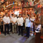 Fotografía Del Gobernador Javier May Rodríguez, Acompañado Por La Flor Tabasco 2026 Y Alcaldes, Durante El Recorrido Por Los Pabellones Municipales En La Nave 2 &Quot;Macuilís&Quot;. El Grupo Se Encuentra Frente A Un Stand Decorado Con Artesanías De Mutusay Y Fibras Naturales, Mientras Un Edil Explica Las Muestras Culturales.