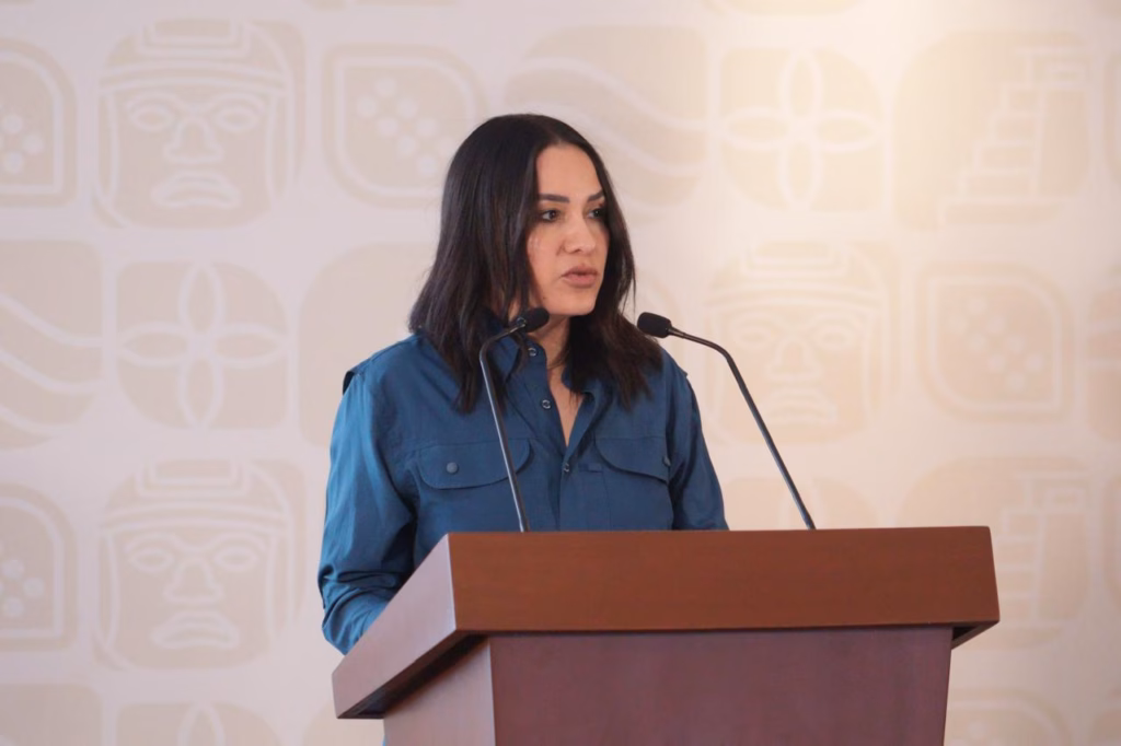 Fotografía De Medio Cuerpo De La Secretaria De Finanzas De Tabasco, Talina Ferrer, Hablando Desde Un Podio De Madera Marrón. Viste Una Camisa De Color Azul Petróleo. De Fondo Se Observa Una Pared Con Logotipos Institucionales Del Gobierno De Tabasco En Tonos Claros. La Funcionaria Tiene Una Expresión Seria Y Profesional Mientras Se Dirige A La Audiencia.