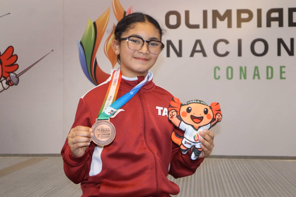 Retrato De La Atleta Tabasqueña Nicole Alexandra Burgos Arrazate Posando Con Su Medalla De Bronce En La Olimpiada Nacional Conade 2026. Viste La Chamarra Deportiva Oficial De La Delegación De Tabasco En Color Guinda Con Detalles Blancos. En Su Mano Derecha Sostiene La Medalla Y En La Izquierda Un Peluche Conmemorativo Del Personaje &Quot;Tlaloque&Quot;. De Fondo Se Observa Un Muro Institucional Con Los Logotipos De La Conade Y El Programa Gabinete Del Agua..