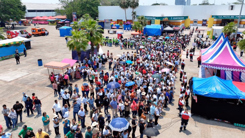 Vista Aérea De Una Explanada En El Parque Tabasco Durante La Feria 2026. Se Observa A Cientos De Personas Formadas De Manera Organizada En Puntos De Reunión Y Rutas De Evacuación Frente A Las Naves De Exposición. Personal De Protección Civil Con Uniformes Rojos Y Blancos Coordina El Flujo De Ciudadanos Bajo El Sol.