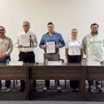 Texto Alternativo (Descripción Para Accesibilidad): Una Fotografía De Interior Con Plano Medio Y Toma Frontal, Bien Iluminada. Cinco Personas Están De Pie En Una Línea Recta Detrás De Una Mesa De Conferencias De Madera Oscura, Posando Para La Cámara. Las Tres Personas En El Centro Sostienen Carpetas Abiertas Mostrando Documentos Firmados. De Izquierda A Derecha: Un Hombre Con Camisa Blanca Y Pantalones De Mezclilla; Un Hombre Mayor Con Camisa De Vestir Blanca Y Pantalones Oscuros; Un Hombre Más Joven En El Centro Con Camisa Azul Abotonada; Una Mujer Con Playera Polo Blanca Y Pantalones Oscuros; Y Un Hombre Con Camisa Verde Claro Abotonada Y Pantalones Oscuros. Sobre La Mesa Hay Tres Botellas De Agua De Plástico Y Cuatro Placas De Identificación Con Texto Apenas Visible. El Fondo Es Una Pared Blanca Y Lisa.