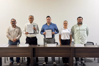Texto Alternativo (Descripción Para Accesibilidad): Una Fotografía De Interior Con Plano Medio Y Toma Frontal, Bien Iluminada. Cinco Personas Están De Pie En Una Línea Recta Detrás De Una Mesa De Conferencias De Madera Oscura, Posando Para La Cámara. Las Tres Personas En El Centro Sostienen Carpetas Abiertas Mostrando Documentos Firmados. De Izquierda A Derecha: Un Hombre Con Camisa Blanca Y Pantalones De Mezclilla; Un Hombre Mayor Con Camisa De Vestir Blanca Y Pantalones Oscuros; Un Hombre Más Joven En El Centro Con Camisa Azul Abotonada; Una Mujer Con Playera Polo Blanca Y Pantalones Oscuros; Y Un Hombre Con Camisa Verde Claro Abotonada Y Pantalones Oscuros. Sobre La Mesa Hay Tres Botellas De Agua De Plástico Y Cuatro Placas De Identificación Con Texto Apenas Visible. El Fondo Es Una Pared Blanca Y Lisa.