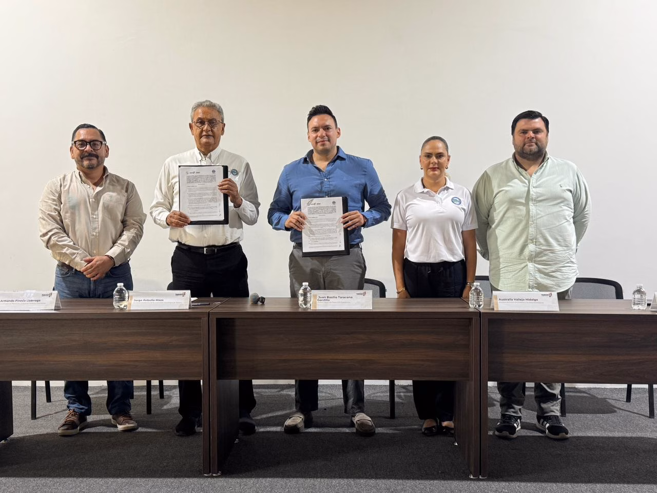 Texto Alternativo (Descripción Para Accesibilidad): Una Fotografía De Interior Con Plano Medio Y Toma Frontal, Bien Iluminada. Cinco Personas Están De Pie En Una Línea Recta Detrás De Una Mesa De Conferencias De Madera Oscura, Posando Para La Cámara. Las Tres Personas En El Centro Sostienen Carpetas Abiertas Mostrando Documentos Firmados. De Izquierda A Derecha: Un Hombre Con Camisa Blanca Y Pantalones De Mezclilla; Un Hombre Mayor Con Camisa De Vestir Blanca Y Pantalones Oscuros; Un Hombre Más Joven En El Centro Con Camisa Azul Abotonada; Una Mujer Con Playera Polo Blanca Y Pantalones Oscuros; Y Un Hombre Con Camisa Verde Claro Abotonada Y Pantalones Oscuros. Sobre La Mesa Hay Tres Botellas De Agua De Plástico Y Cuatro Placas De Identificación Con Texto Apenas Visible. El Fondo Es Una Pared Blanca Y Lisa.