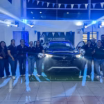 Una Fotografía De Ángulo Medio Captura A Un Grupo Grande De Doce Empleados, De Diversas Edades Y Géneros, Posando Con Entusiasmo En Un Concesionario De Volkswagen Para El Lanzamiento De Una Edición Limitada. Visten Camisetas De Uniforme Negras Con Logos De Volkswagen Y Goal 2026.