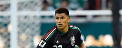 El Futbolista Mexicano Jesús Gallardo En Plena Acción Sobre El Césped Durante Un Partido De Fútbol. Viste El Uniforme Negro De La Selección Mexicana Con El Número 23 En El Pecho Y En El Short, Detalles Tricolores En Los Hombros, Calcetas Negras Altas Y Tacos De Color Rosa Brillante. Su Mirada Está Fija Al Frente, Manteniendo Una Postura Alerta Cerca Del Balón, El Cual Se Observa En El Pasto A La Esquina Inferior Derecha. Al Fondo Desenfocado Se Aprecian Las Tribunas Y Una Portería.