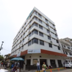Fotografía En Contrapicado De Un Edificio De Varios Pisos Con Fachada Blanca Y Ventanas Oscuras, Ubicado En Una Esquina Urbana. En La Planta Baja Se Observa Un Letrero Que Identifica Las Instalaciones De La Villa Universitaria, Junto Con Logotipos Institucionales Del Gobierno De Tabasco. Varias Personas Transitan Por La Acera Y Se Aprecian Vehículos Estacionados A Lo Largo De La Calle Lateral Bajo Un Cielo Nublado, Capturando La Infraestructura Del Complejo Residencial.