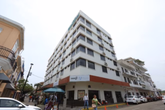 Fotografía En Contrapicado De Un Edificio De Varios Pisos Con Fachada Blanca Y Ventanas Oscuras, Ubicado En Una Esquina Urbana. En La Planta Baja Se Observa Un Letrero Que Identifica Las Instalaciones De La Villa Universitaria, Junto Con Logotipos Institucionales Del Gobierno De Tabasco. Varias Personas Transitan Por La Acera Y Se Aprecian Vehículos Estacionados A Lo Largo De La Calle Lateral Bajo Un Cielo Nublado, Capturando La Infraestructura Del Complejo Residencial.