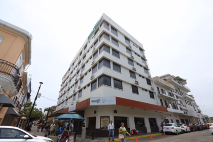 Fotografía En Contrapicado De Un Edificio De Varios Pisos Con Fachada Blanca Y Ventanas Oscuras, Ubicado En Una Esquina Urbana. En La Planta Baja Se Observa Un Letrero Que Identifica Las Instalaciones De La Villa Universitaria, Junto Con Logotipos Institucionales Del Gobierno De Tabasco. Varias Personas Transitan Por La Acera Y Se Aprecian Vehículos Estacionados A Lo Largo De La Calle Lateral Bajo Un Cielo Nublado, Capturando La Infraestructura Del Complejo Residencial.