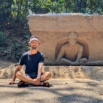 Un Plano Medio Completo Muestra Al Creador De Contenido Omar Nok Sentado Con Las Piernas Cruzadas Sobre Un Sendero Pavimentado En El Parque Museo La Venta. Viste Una Playera Negra, Shorts, Sandalias Y Un Sombrero De Pescador Color Gris Claro. Detrás De Él, Imita La Postura De Una Escultura Olmeca Tallada En Un Gran Altar De Piedra Rectangular. La Figura Prehispánica Emerge De Un Nicho En Posición Sedente Y Con Las Piernas Cruzadas. El Entorno Está Rodeado De Abundante Vegetación Selvática Y Árboles, Con La Luz Del Sol Filtrándose Entre Las Hojas Y Creando Sombras En El Suelo.