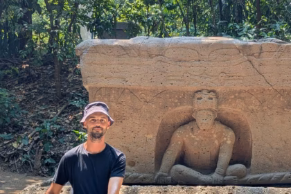 Un Plano Medio Completo Muestra Al Creador De Contenido Omar Nok Sentado Con Las Piernas Cruzadas Sobre Un Sendero Pavimentado En El Parque Museo La Venta. Viste Una Playera Negra, Shorts, Sandalias Y Un Sombrero De Pescador Color Gris Claro. Detrás De Él, Imita La Postura De Una Escultura Olmeca Tallada En Un Gran Altar De Piedra Rectangular. La Figura Prehispánica Emerge De Un Nicho En Posición Sedente Y Con Las Piernas Cruzadas. El Entorno Está Rodeado De Abundante Vegetación Selvática Y Árboles, Con La Luz Del Sol Filtrándose Entre Las Hojas Y Creando Sombras En El Suelo.