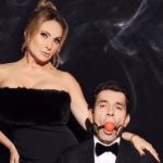 Retrato Promocional De Los Actores Elizabeth Álvarez Y Jorge Salinas Sobre Un Fondo Negro Con Sutiles Ráfagas De Humo Blanco. A La Izquierda, La Actriz Posa De Pie Con Un Vestido Negro Strapless Texturizado, Pendientes Largos Y El Cabello Recogido, Mirando Fijamente A La Cámara Con Una Expresión De Poder. A La Derecha, En Primer Plano, El Actor Viste Un Esmoquin Negro Con Pajarita Y Aparece Amordazado Con Una Mordaza De Bola Roja Sujeta Por Correas De Cuero Negras, Mientras La Mano De La Actriz Reposa Sobre Su Hombro.