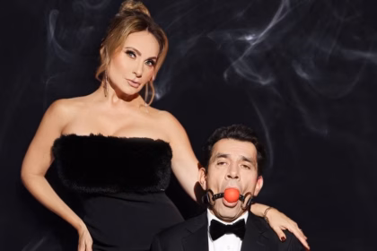 Retrato Promocional De Los Actores Elizabeth Álvarez Y Jorge Salinas Sobre Un Fondo Negro Con Sutiles Ráfagas De Humo Blanco. A La Izquierda, La Actriz Posa De Pie Con Un Vestido Negro Strapless Texturizado, Pendientes Largos Y El Cabello Recogido, Mirando Fijamente A La Cámara Con Una Expresión De Poder. A La Derecha, En Primer Plano, El Actor Viste Un Esmoquin Negro Con Pajarita Y Aparece Amordazado Con Una Mordaza De Bola Roja Sujeta Por Correas De Cuero Negras, Mientras La Mano De La Actriz Reposa Sobre Su Hombro.