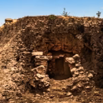 Fotografía En Plano General Que Muestra Un Montículo Arqueológico De Tierra Y Piedras Excavado En Un Entorno Semiárido Con Vegetación Baja, Como Agaves A La Izquierda. Al Centro Del Montículo Se Abre Una Estructura De Piedra Colapsada Que Revela El Acceso A Una Cripta O Cámara Fúnebre Subterránea. El Paisaje De Fondo Incluye Colinas, Valles Verdes Bajo Un Cielo Azul Despejado Con Nubes Ligeras, Y Restos De Muros De Piedra A La Derecha.