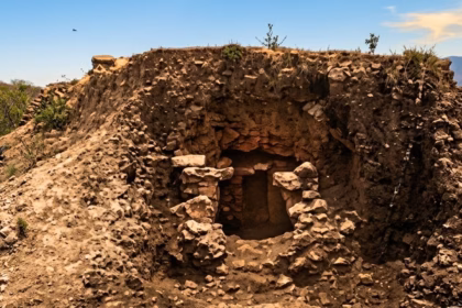 Fotografía En Plano General Que Muestra Un Montículo Arqueológico De Tierra Y Piedras Excavado En Un Entorno Semiárido Con Vegetación Baja, Como Agaves A La Izquierda. Al Centro Del Montículo Se Abre Una Estructura De Piedra Colapsada Que Revela El Acceso A Una Cripta O Cámara Fúnebre Subterránea. El Paisaje De Fondo Incluye Colinas, Valles Verdes Bajo Un Cielo Azul Despejado Con Nubes Ligeras, Y Restos De Muros De Piedra A La Derecha.