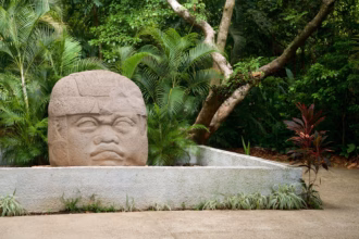 Una Cabeza Colosal Olmeca De Piedra Exhibida Al Aire Libre En El Parque Museo La Venta, Rodeada De Abundante Vegetación Tropical, Palmeras, Árboles Y Un Sendero Pavimentado Delimitado Por Una Jardinera De Concreto Gris.