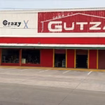 Fotografía De La Fachada Exterior De Una Sucursal De La Tienda Casa Gutza, Vista Desde El Otro Lado De La Calle. El Edificio Tiene Una Amplia Planta Baja Pintada De Rojo Brillante Con Columnas Delineadas En Color Amarillo, Y Una Sección Superior De Lámina Blanca Donde Se Ubican Los Anuncios Comerciales. En El Centro Destaca El Logotipo Principal De 'Casa Gutza'.