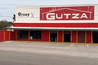 Fotografía De La Fachada Exterior De Una Sucursal De La Tienda Casa Gutza, Vista Desde El Otro Lado De La Calle. El Edificio Tiene Una Amplia Planta Baja Pintada De Rojo Brillante Con Columnas Delineadas En Color Amarillo, Y Una Sección Superior De Lámina Blanca Donde Se Ubican Los Anuncios Comerciales. En El Centro Destaca El Logotipo Principal De 'Casa Gutza'.