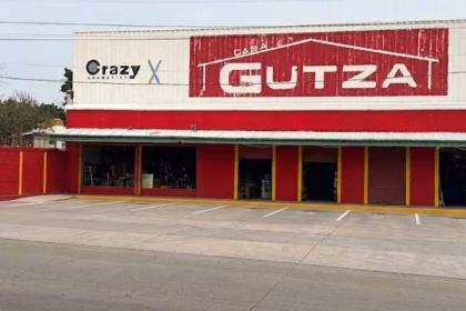 Fotografía De La Fachada Exterior De Una Sucursal De La Tienda Casa Gutza, Vista Desde El Otro Lado De La Calle. El Edificio Tiene Una Amplia Planta Baja Pintada De Rojo Brillante Con Columnas Delineadas En Color Amarillo, Y Una Sección Superior De Lámina Blanca Donde Se Ubican Los Anuncios Comerciales. En El Centro Destaca El Logotipo Principal De 'Casa Gutza'.